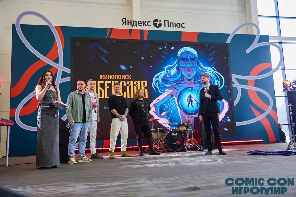 На изображении представлены люди на мероприятии с сценой и баннером. Сцена украшена графикой, возможно, относящейся к комиксам или играм.