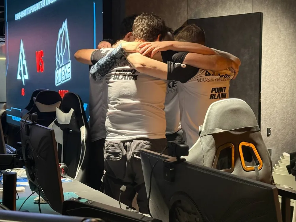 Группа людей обнимается, вероятно, после игры. На заднем плане видны игровые компьютеры и экран с логотипами команд.