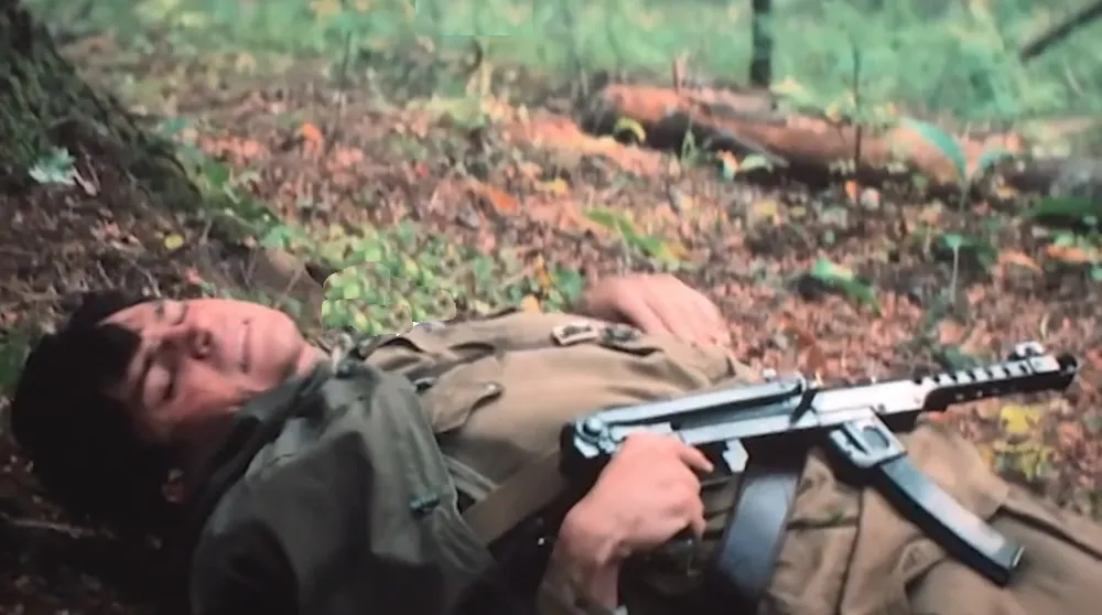 Человек в военной форме лежит на земле с автоматом в лесной местности.