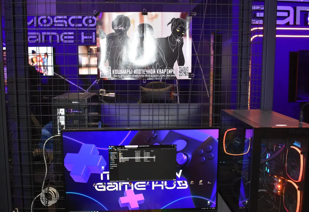 Изображение игрового пространства с монитором, оформленным в темных тонах и неоновыми элементами. На стене висит постер с изображением персонажей.