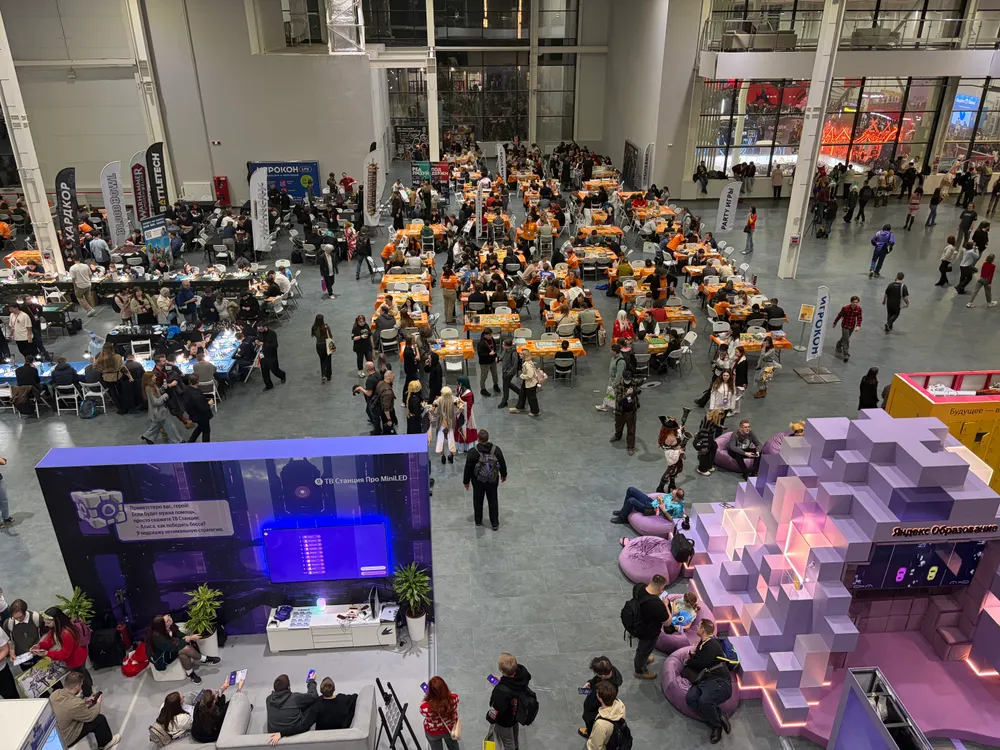 Изображение большого количества людей в помещении, вероятно, на мероприятии или выставке. Видны столы и зоны для отдыха.