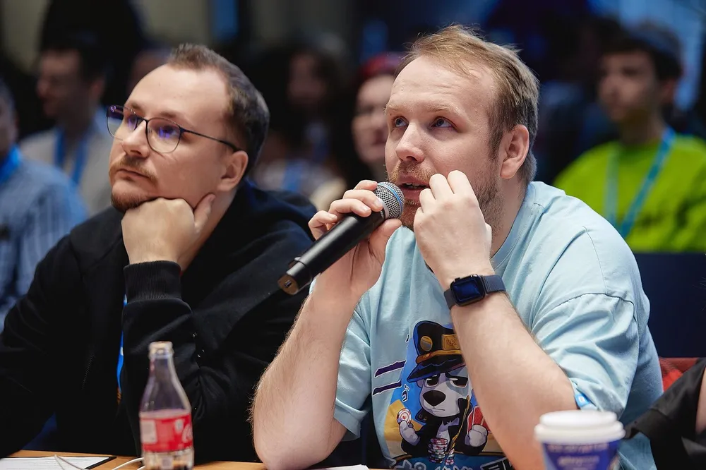 Два человека на мероприятии, один из них держит микрофон и задаёт вопрос. На заднем плане видны зрители.