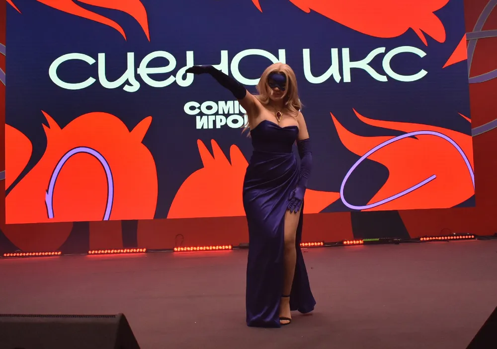 На изображении женщина в вечернем платье, позирующая на сцене. Фон с текстом, вероятно, связанным с шоу или мероприятием.
