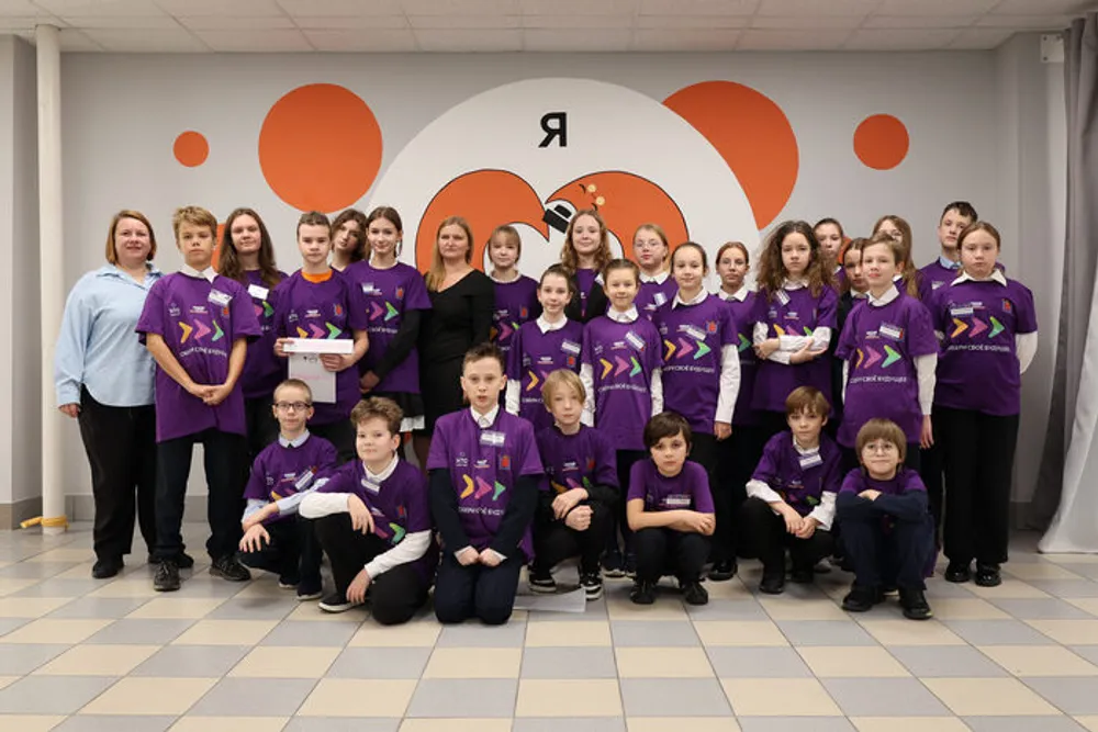 Группа детей и взрослых в одинаковых фиолетовых футболках, позирующая на фоне стены с оранжевыми элементами. Сцена из мероприятия или праздника.