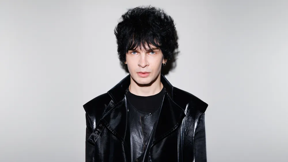 Человек в черной одежде с короткой прической на нейтральном фоне.