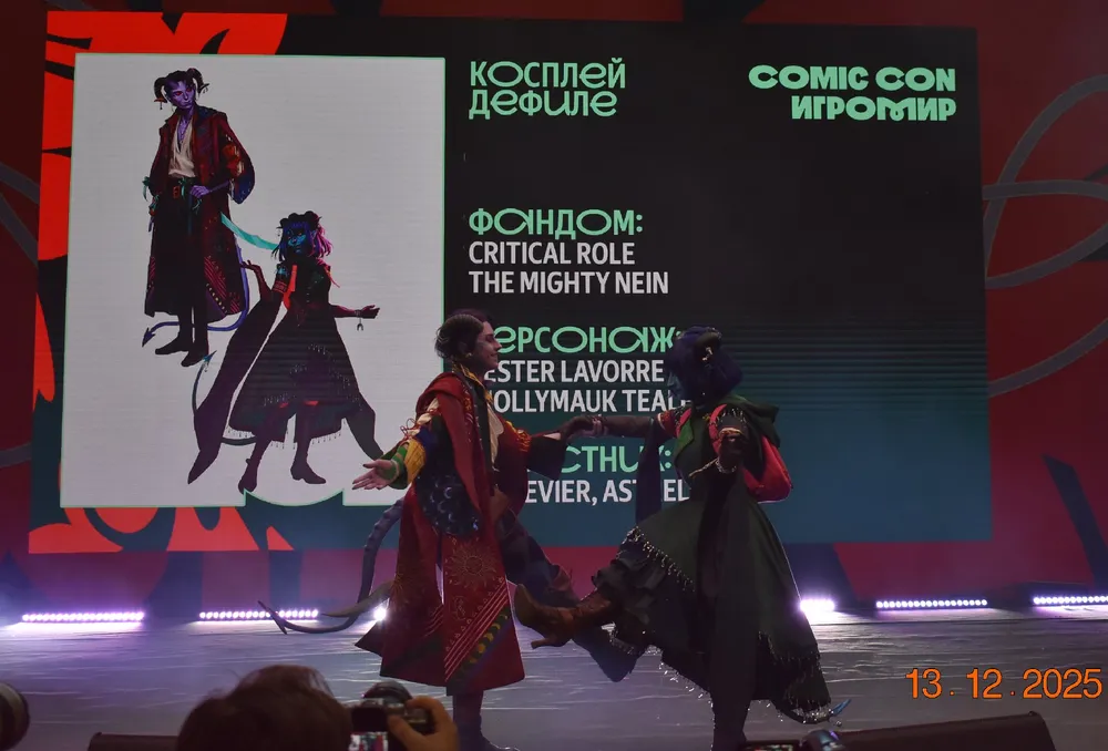 Косплей на мероприятии Comic Con, персонажи с необычными костюмами, сцена и экран с информацией о мероприятии.