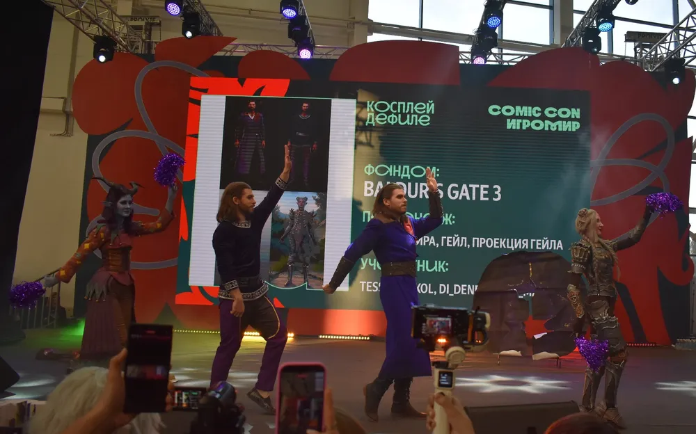 На изображении видно сцену с выступлением танцовщиц на мероприятии Comic Con, декорации и экран с надписью "Baldur\