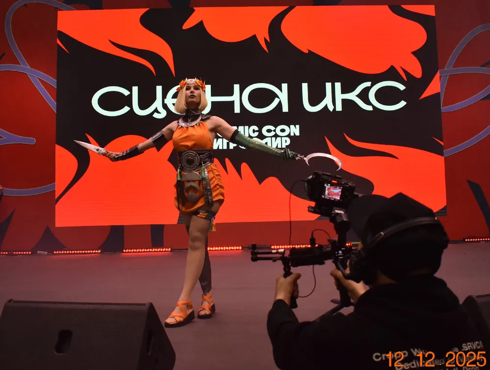 Изображение человека в костюме на сцене мероприятия, с камерой оператора и фоном с надписями.