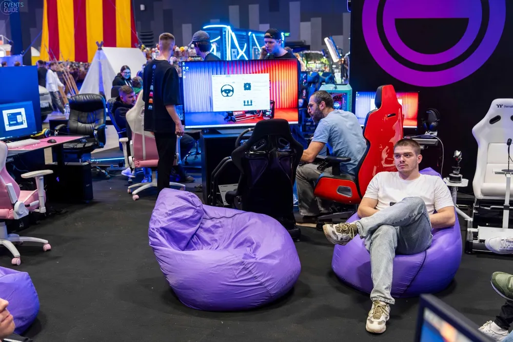 Изображение людей в игровом пространстве, сидящих на пуфах, с компьютерами и яркими экранами на заднем плане.