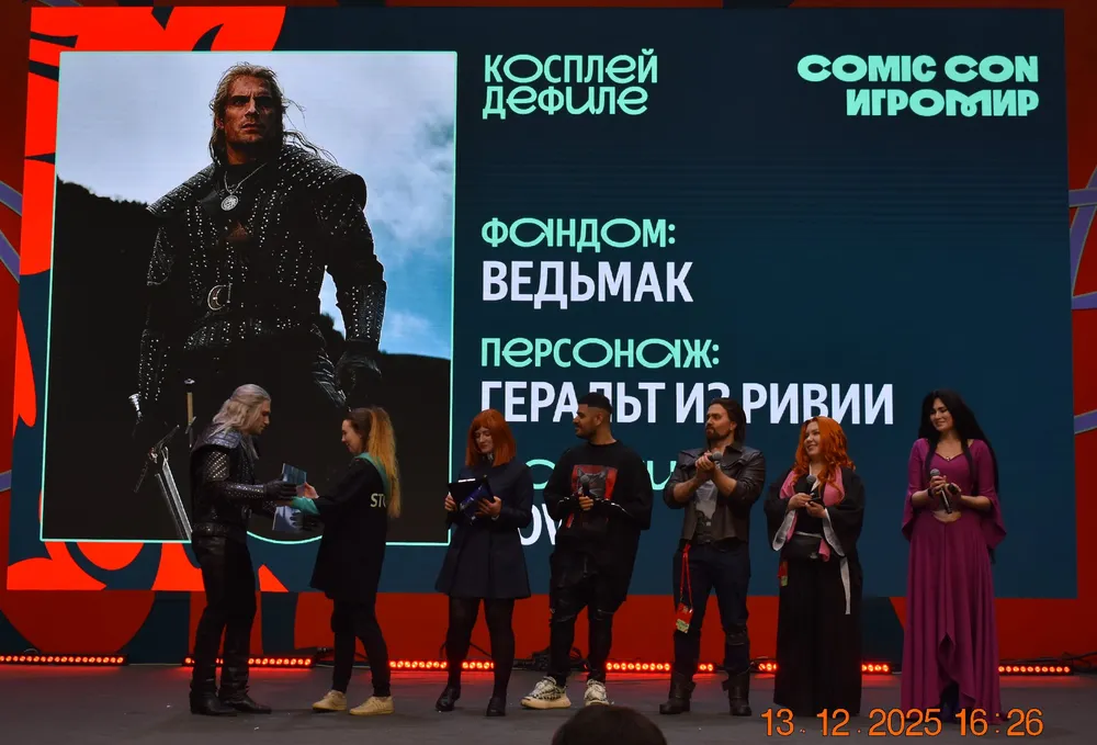 Сцена с косплеем на мероприятии Comic Con. На сцене персонаж и участники в костюмах. На экране указаны название фандома \
