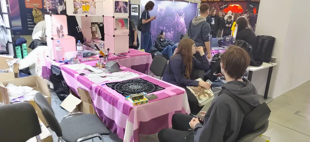 На изображении показан выставочный стенд с несколькими людьми, работающими и общающимися. Присутствуют столы с различными предметами, в том числе с розовой скатертью.