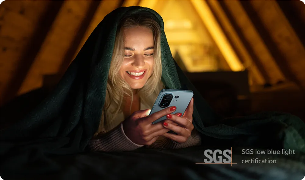 На изображении девушка, укрытая одеялом, улыбается и смотрит на телефон. Фон деревянной конструкции.