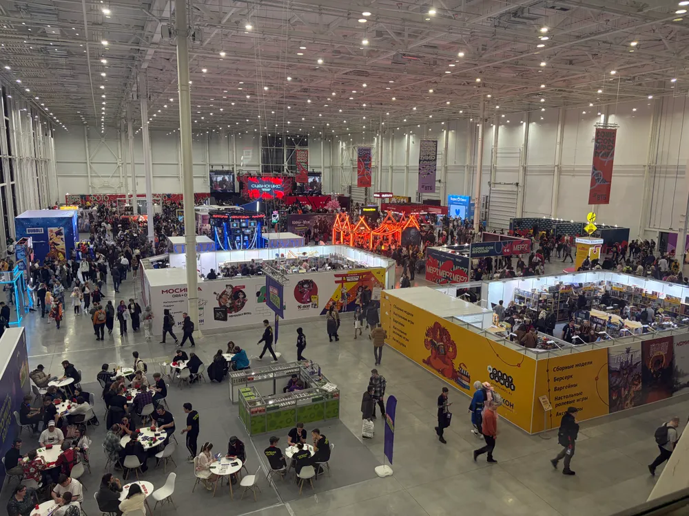 Изображение представляет собой большое помещение с людьми, участниками мероприятия, торговыми стендами и выставочными зоной. Видно много столов и стульев, а также рекламные баннеры.