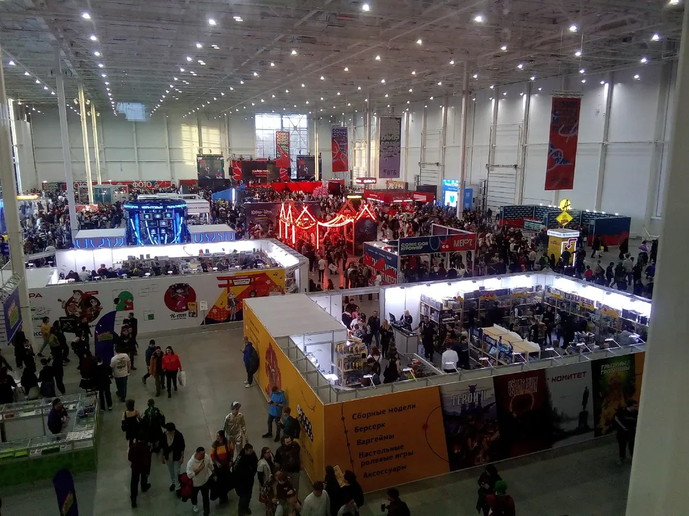 Изображение большого выставочного зала с множеством людей и стендами. Многочисленные выставочные стенды, освещение, общий вид мероприятия.