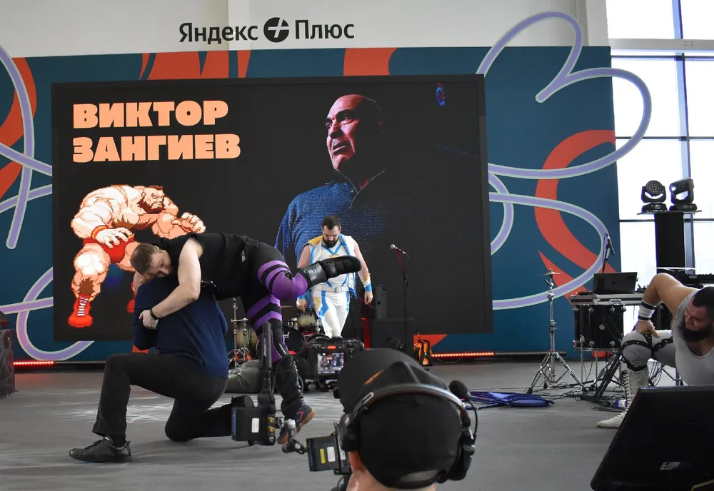 На изображении представлены люди, занимающиеся физической активностью на фоне большого экрана с изображением мужчины. Сцена выглядит как мероприятие или соревнование.