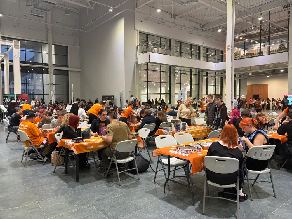 Изображение показывает людей, сидящих за столами в большом помещении, возможно, на мероприятии или собрании.