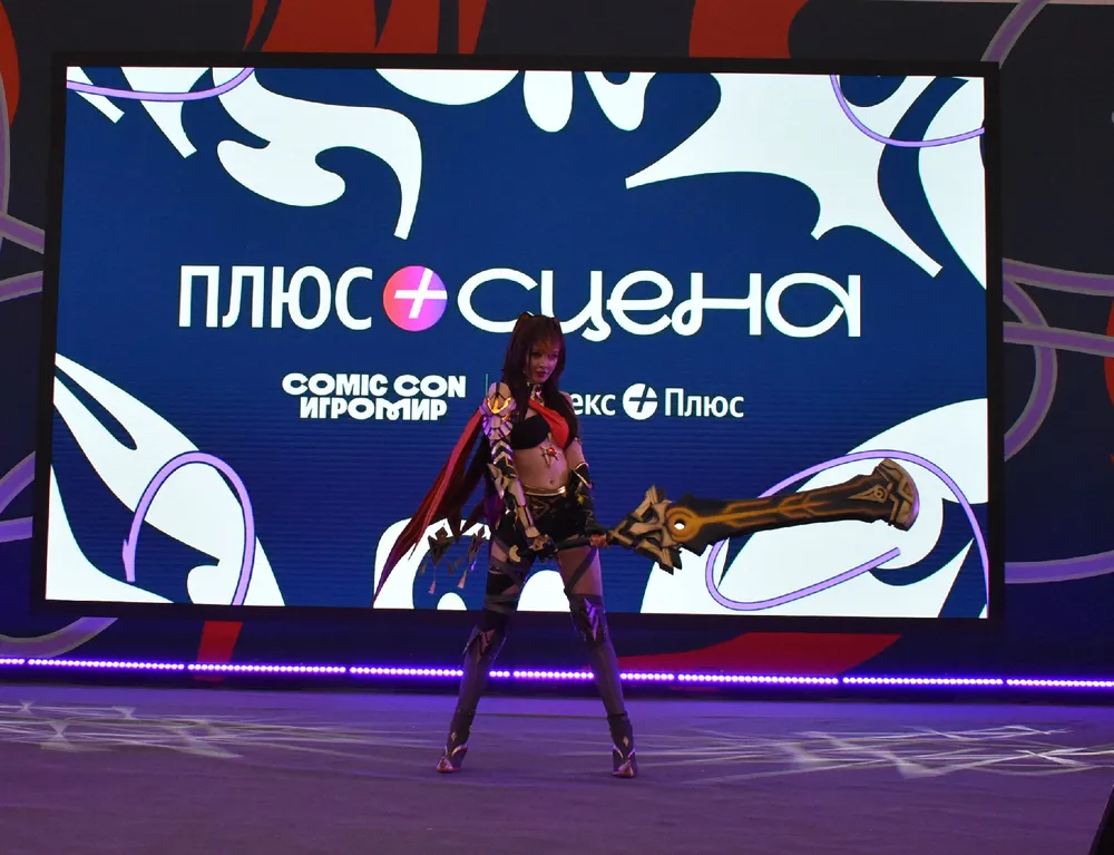 На изображении персонаж в костюме с оружием, фон с логотипом и надписью. Сцена с мероприятия, связанного с комиксами.
