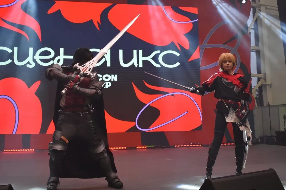 Изображение двух человек в костюмах, выступающих на сцене. Фон с надписью и графическими элементами.