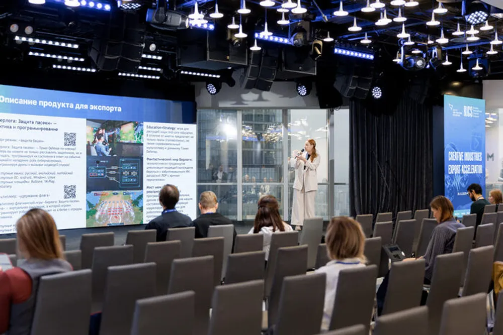 Изображение деловой обстановки, женщина выступает на сцене перед зрителями, на стендах информация, связанная с презентацией.