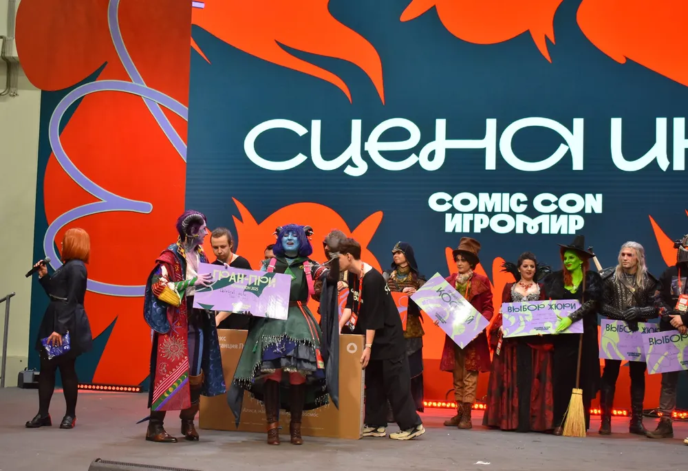 Изображение с людьми на мероприятии, возможно, связанном с комиксами или косплеем. Люди в костюмах, сцена, фон с надписью \