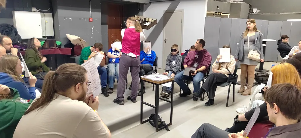 Группа людей, сидящих в помещении, некоторые из них держат бумажные плакаты или таблички.