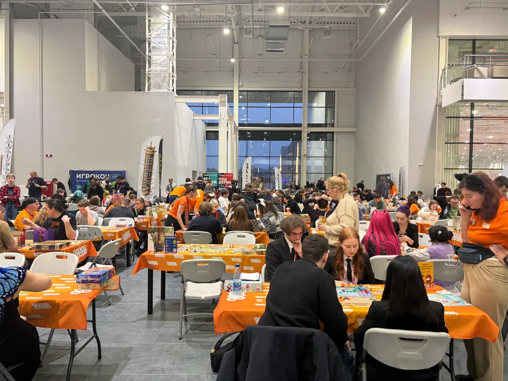 Сцена с людьми, сидящими за столами в большом помещении, вероятно, на мероприятии или выставке.