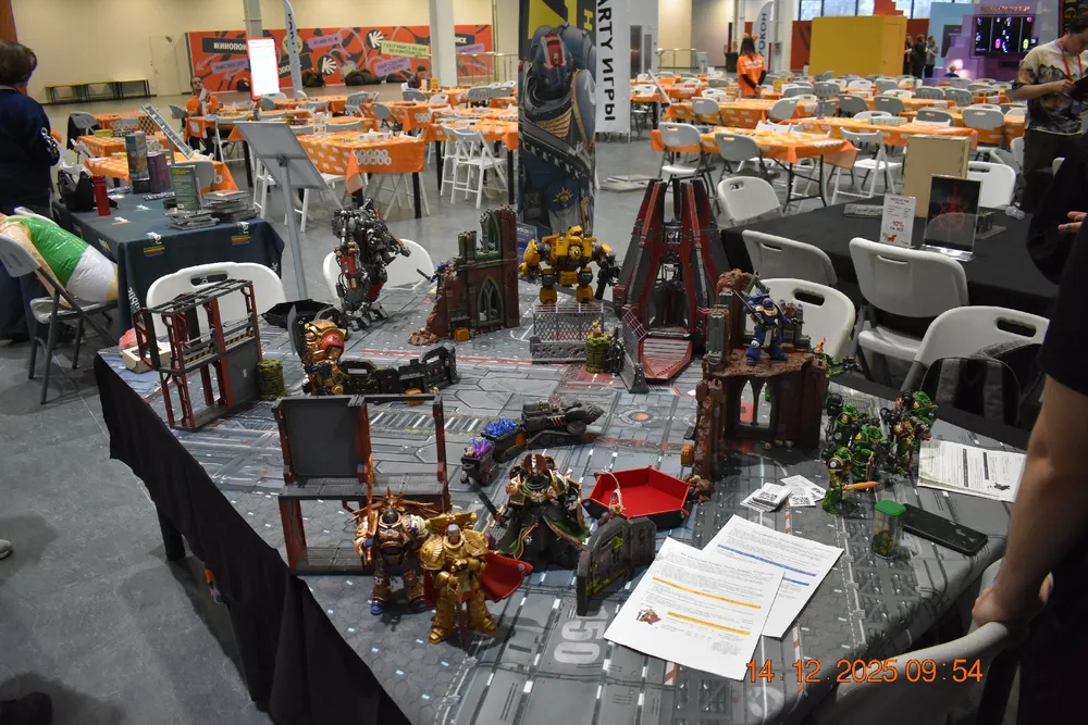 На картинке изображена игровая площадка с миниатюрами и декорациями, а также столы с сиденьями в зале.