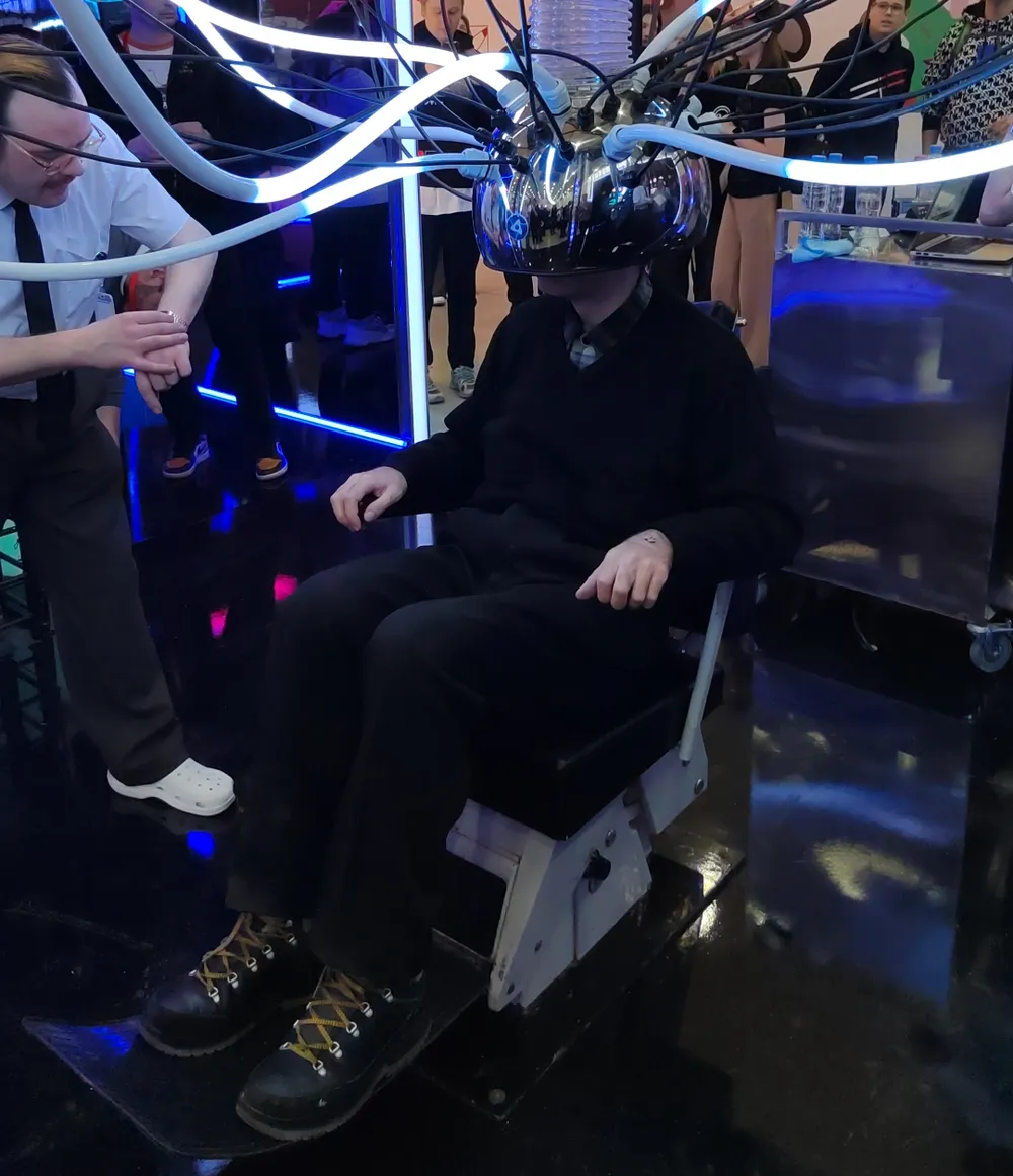 На изображении человек в кресле с необычной аппаратурой на голове, вокруг него другие люди и элементы освещения.