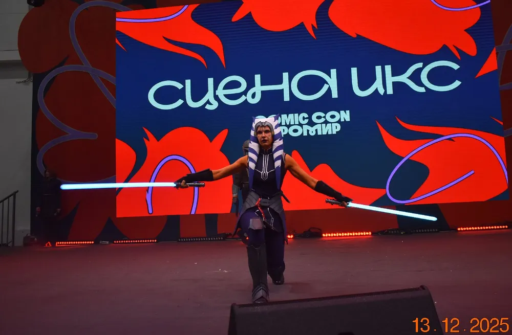 На изображении человек в костюме с световыми мечами, выступающий на сцене с фоном Comic Con.