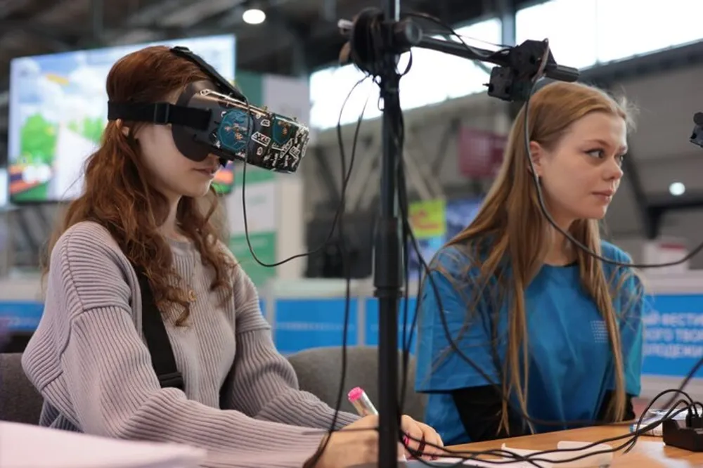 Две девушки, одна из которых носит очки виртуальной реальности, сидят за столом на мероприятии или выставке.