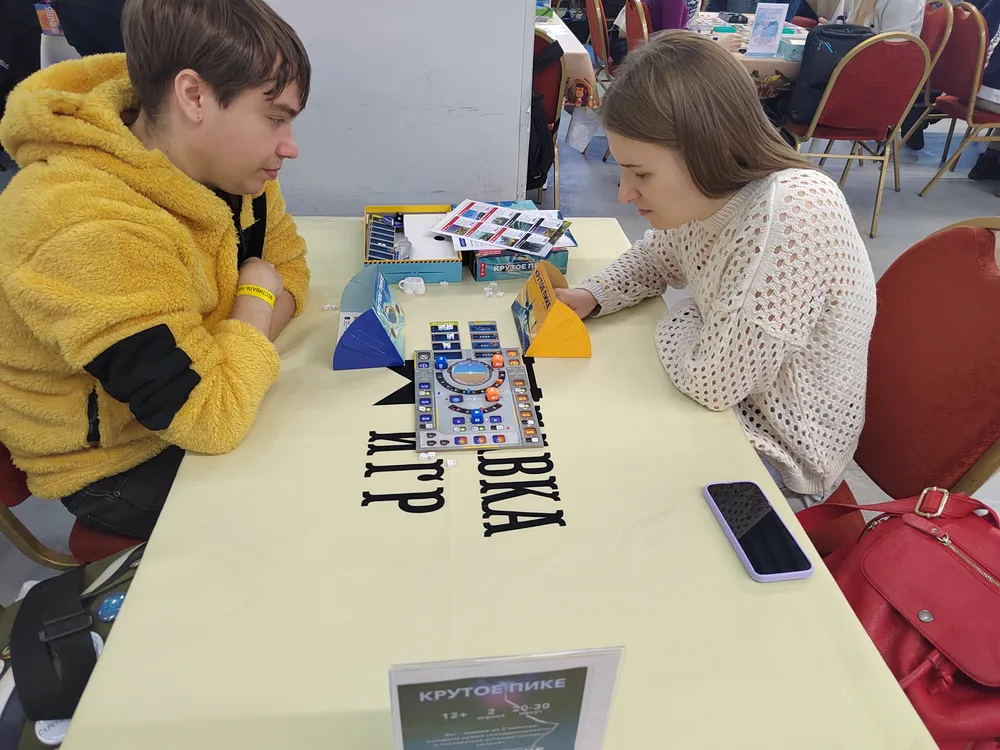 Два человека играют за столом, на котором расположены игровые элементы и телефон. На заднем плане видны стулья.