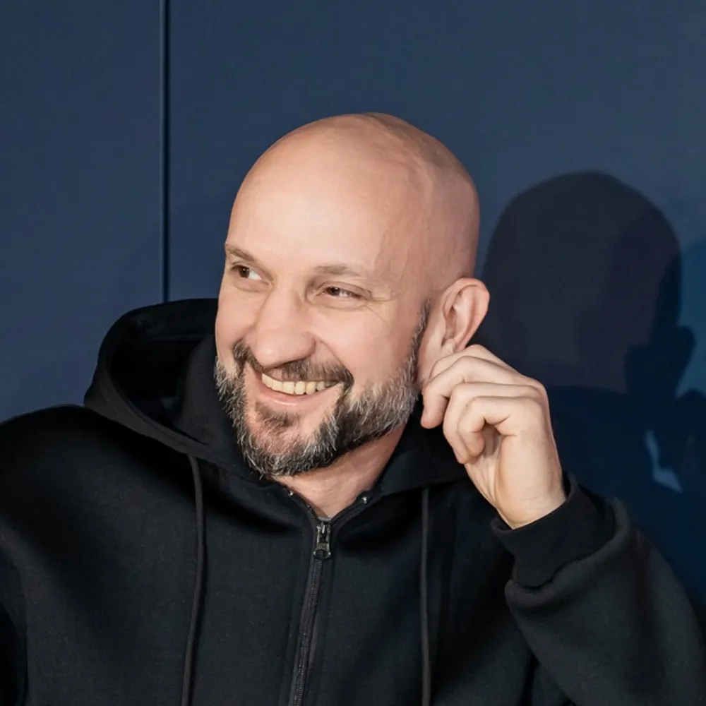 На изображении человек с короткой бородой и лысиной, улыбается, одет в черный худи, фон темно-синий.