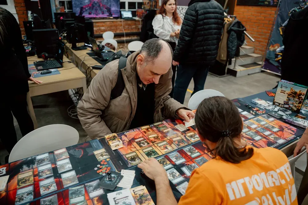 Два человека играют в настольную игру на мероприятии, в фоне другие люди и столы с компьютерной техникой.