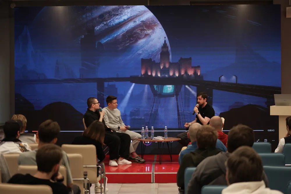 На изображении присутствуют три человека, общающихся на фоне sci-fi пейзажа. Видна аудитория, слушающая их.
