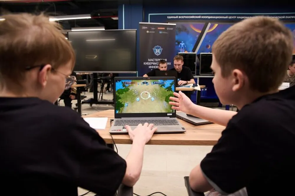 Два мальчика, сидящие за столом, играют на ноутбуке. На фоне видны другие люди и оборудование.