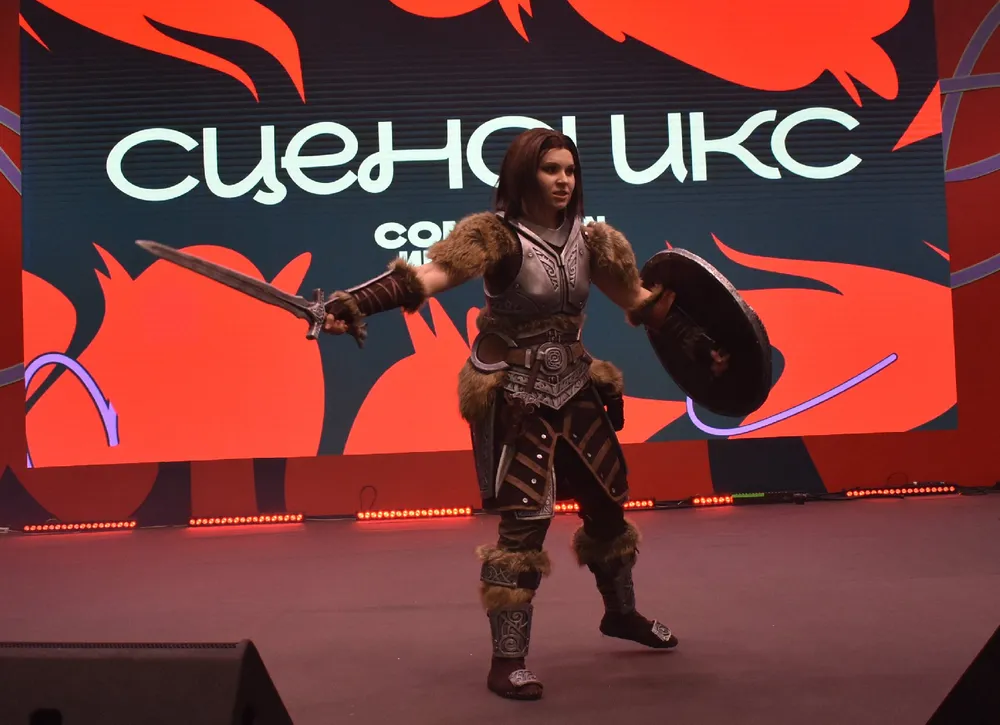 Изображение человека в костюме на мероприятии, с мечом и щитом, на фоне сценического оформления.