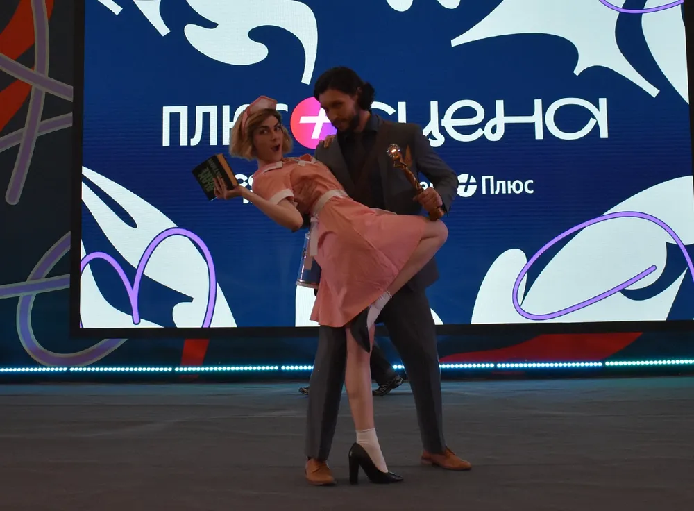 На изображении люди, исполняющие танцевальный номер на сцене, с фоновым баннером.