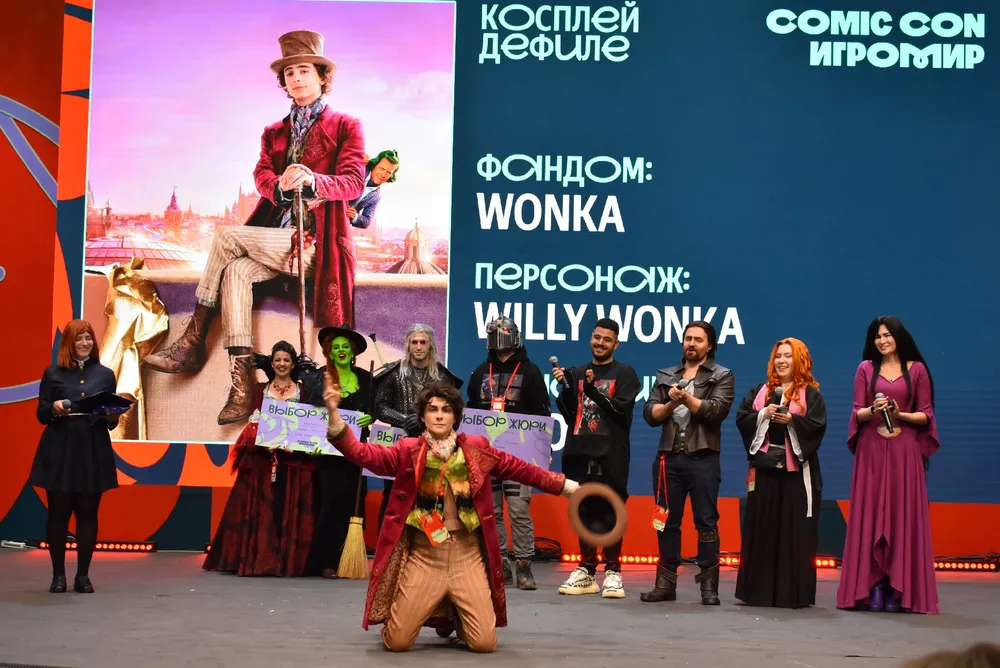 Косплей на мероприятии, участники в костюмах, на сцене, фон с надписями о фэндоме и персонаже.