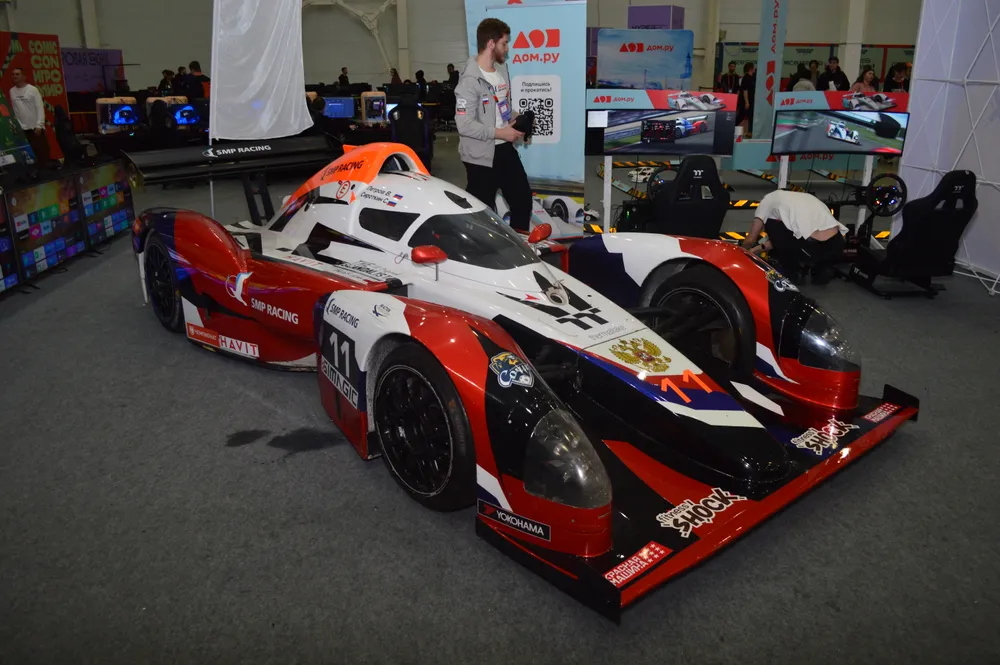 Изображение гоночного автомобиля на выставке, рядом с ним человек.