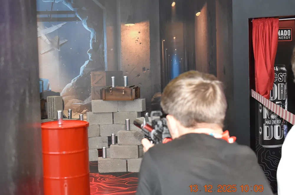 На картинке изображён человек, который целится из игрушечного оружия. Фон включает в себя декорации, такие как стены и бочки.