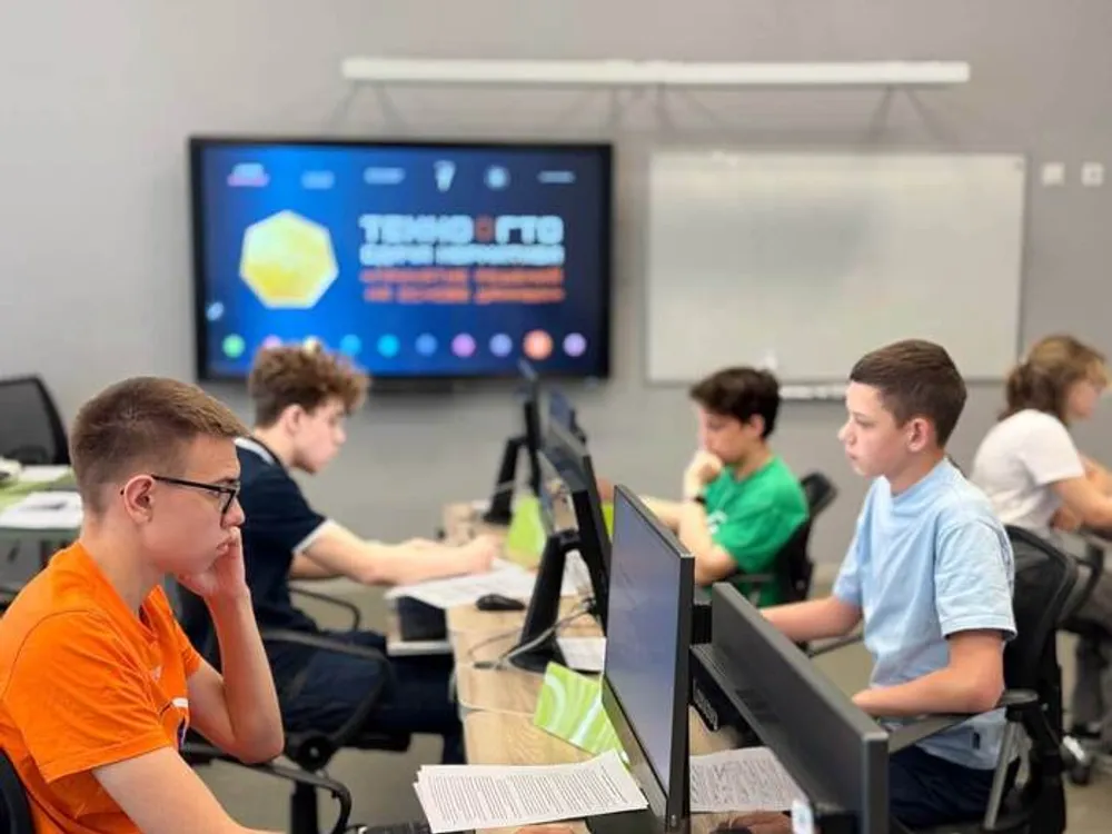 Группа подростков, сидящих за компьютерами в классе. На заднем плане видно экран с надписью.