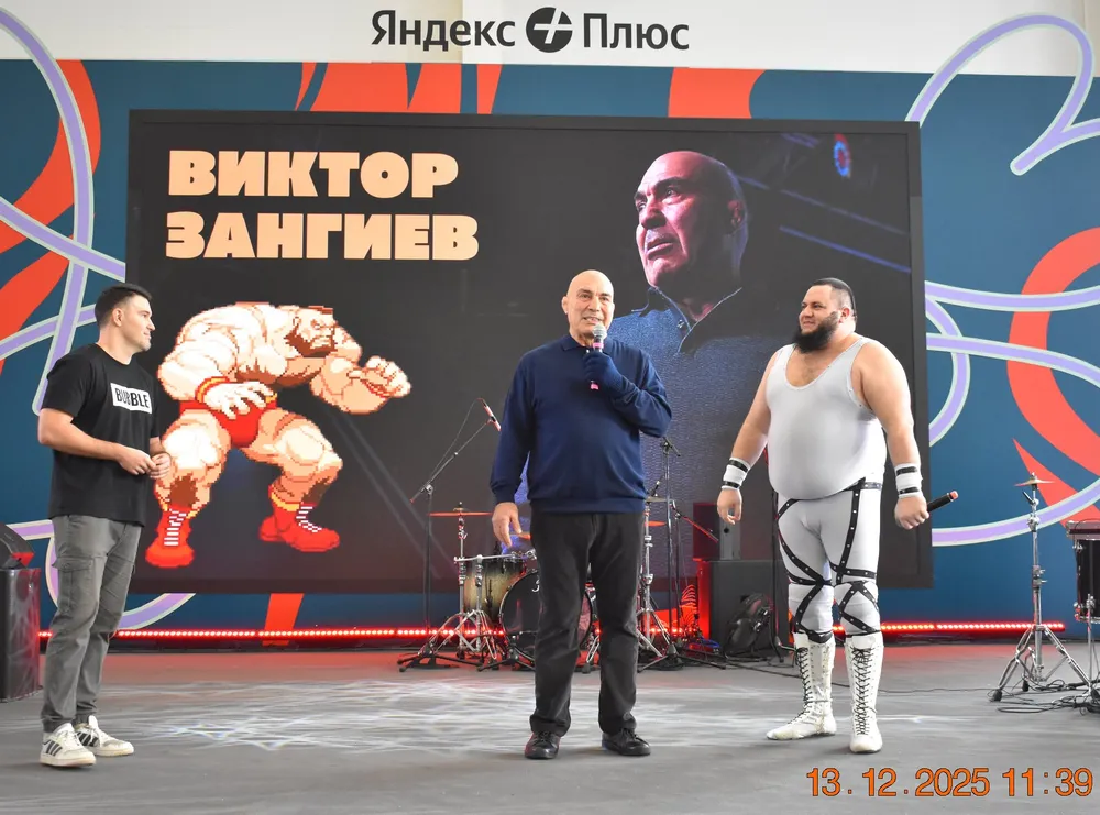 На изображении представлены мужчины на мероприятии, один из них выступает с речью, на заднем плане виден экран с изображением и текстом.