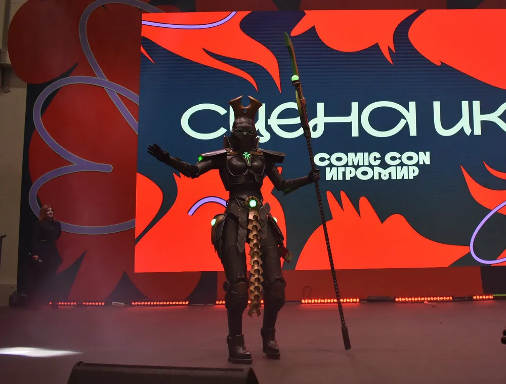 На изображении представлен человек в костюме на сцене, возможно, на мероприятии Comic Con. На фоне виден баннер с надписью \