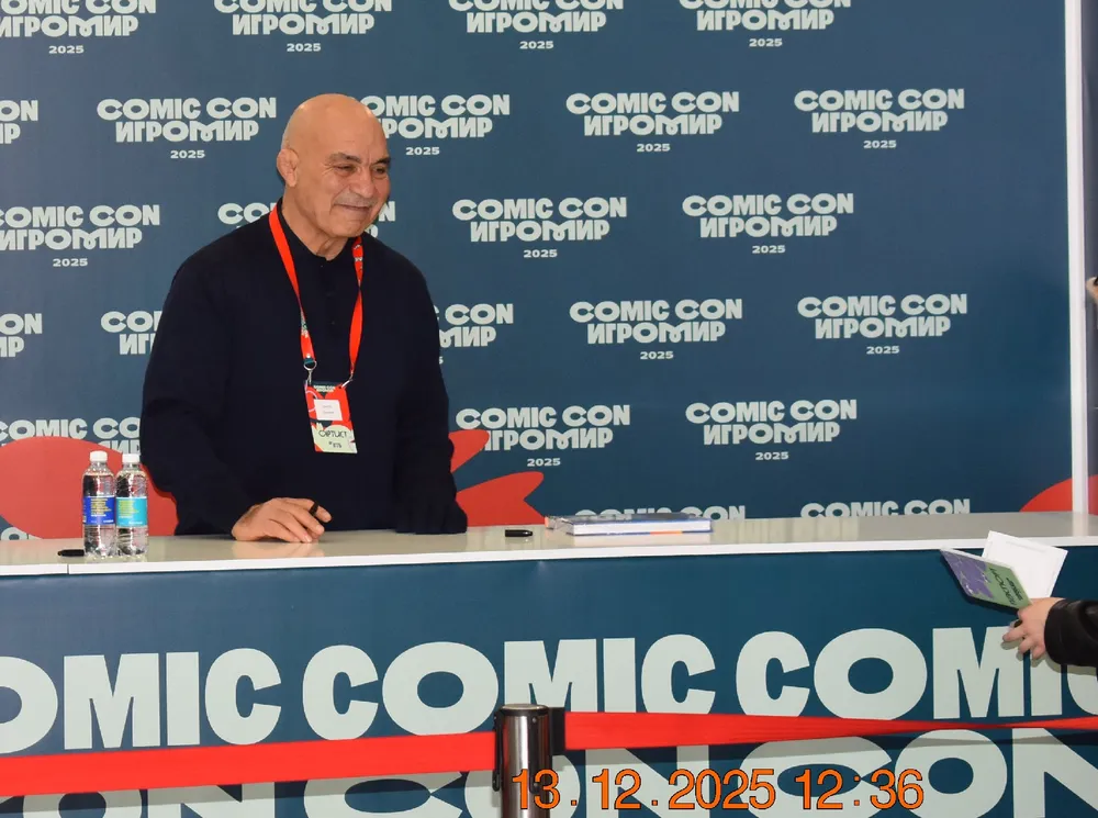 На изображении человек за столом на мероприятии Comic Con, с логотипами и надписями на фоне.