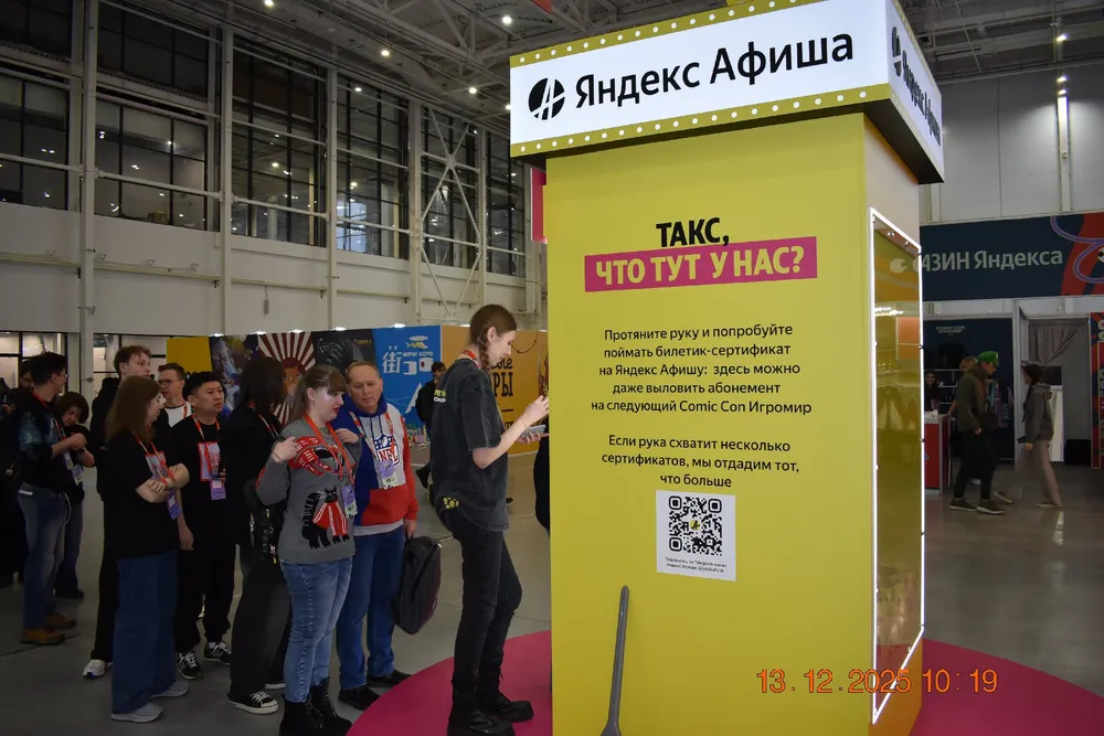 Изображение показывает стенд Яндекс Афиши на мероприятии, люди стоят в очереди, есть QR-код и текст на стенде.