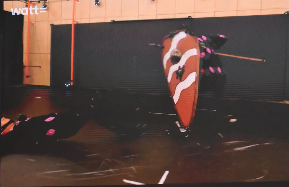 Изображение людей в костюмах, один человек с щитом, действие на арене или сцене.