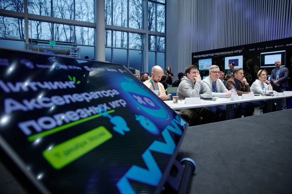 На изображении виден стол с участниками мероприятия, акцент на логотипе и тексте на экране. Вся сцена в помещении с окнами.