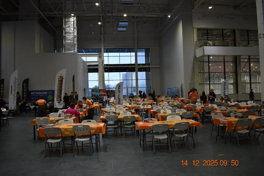 Изображение интерьерного пространства с рядами столов и стульев, присутствуют люди, обстановка выглядит как место для мероприятия.