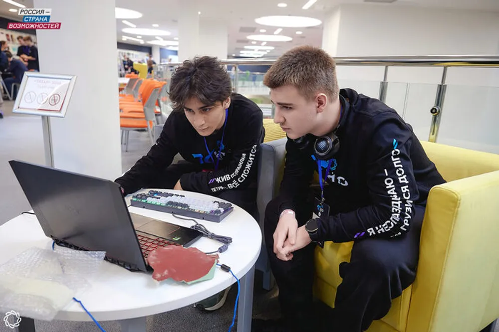 Два человека сидят за столом с ноутбуком, один из них смотрит на экран, другой что-то показывает. Окружение современное, с яркими цветами и мебелью.