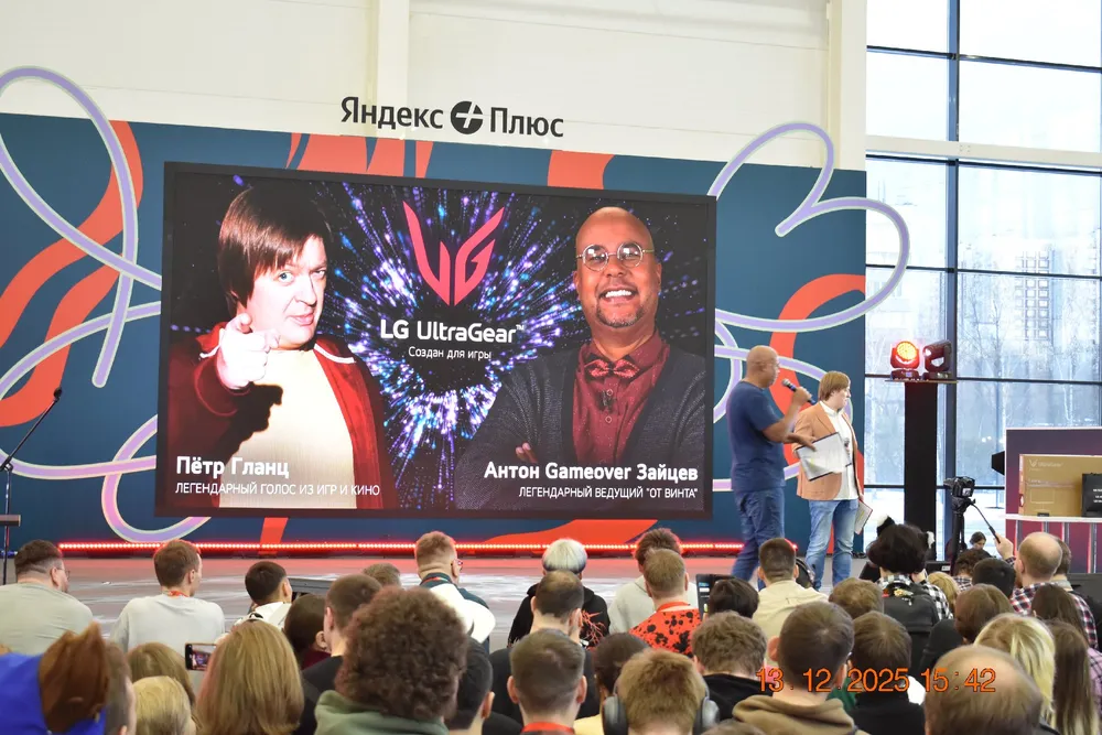 На изображении представлены два человека на сцене с большим экраном, на котором отображены их имена и информация о мероприятии. В зале присутствует аудитория.